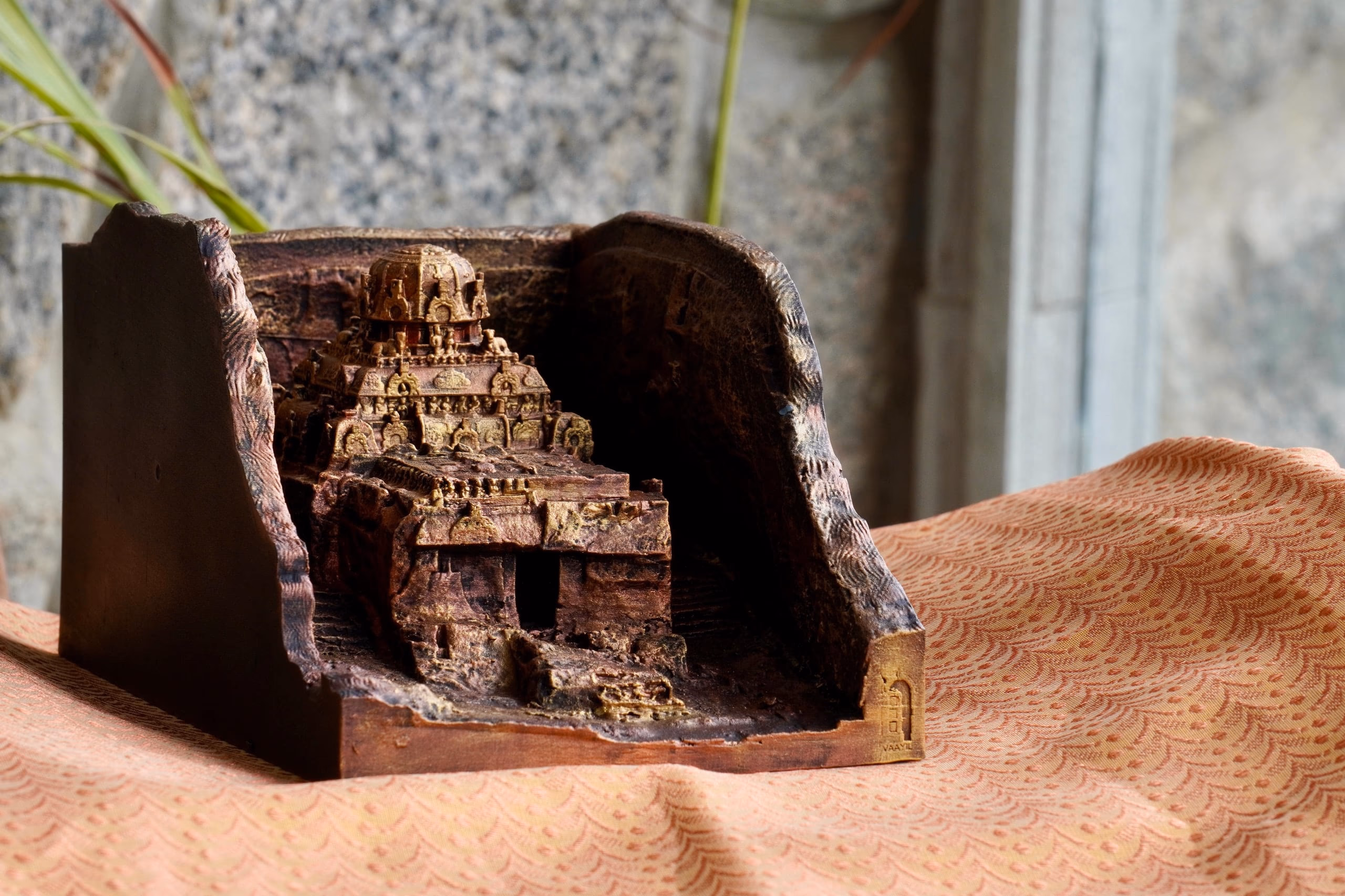 A 3D-printed replica of Vettuvan Koil photographed with its quarried pit, lit from one side on block-printed fabric, with an embossed Vaayil plaque on the base corner.